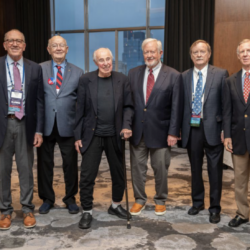 A group of six older adults, all wearing suits, standing in a room with large windows and wooden paneling. One person is using a cane. They are likely attending an event or gathering.